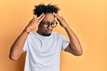 Young african american man with beard wearing casual white t shirt and glasses with hand on head, headache because stress. suffering migraine.