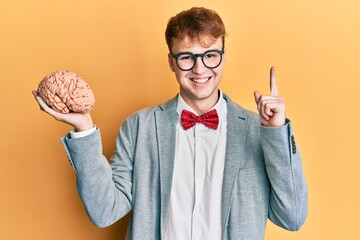 Young caucasian man wearing glasses holding brain smiling with an idea or question pointing finger...