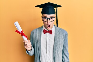 Young caucasian nerd man wearing glasses and graduation cap and holding degree scared and amazed...
