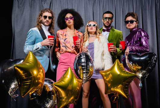 glamorous interracial friends standing near grey curtain on black background.