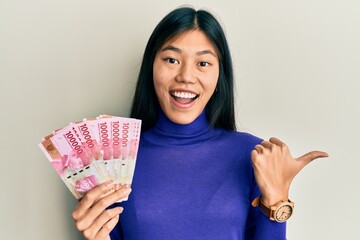 Young chinese woman holding indonesian rupiah banknotes pointing thumb up to the side smiling happy with open mouth