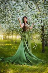 Fototapeta na wymiar A beautiful young woman in a green dress with a large train among the blooming apple trees.