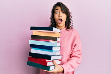 Young hispanic woman holding a pile of books afraid and shocked with surprise and amazed expression, fear and excited face.