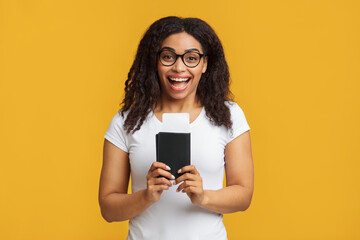 Cheap flights. Overjoyed black lady holding passport and tickets, standing over yellow background