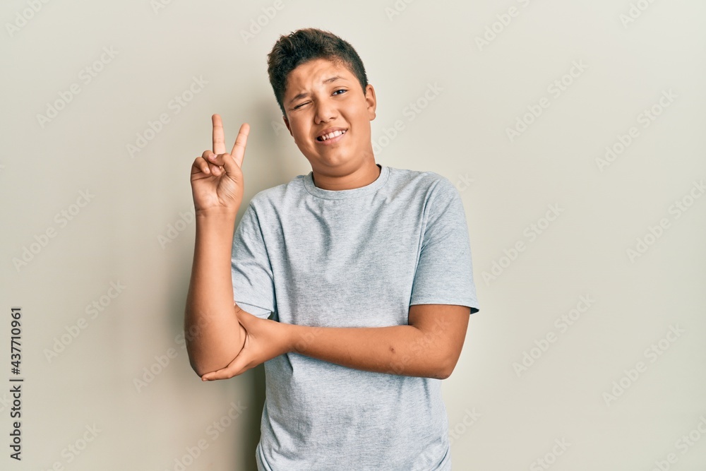 Wall mural Teenager hispanic boy wearing casual grey t shirt smiling with happy face winking at the camera doing victory sign. number two.