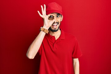 Young hispanic man wearing delivery uniform and cap smiling happy doing ok sign with hand on eye looking through fingers