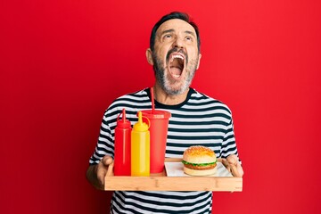 Middle age hispanic man eating a tasty classic burger and soda angry and mad screaming frustrated...