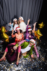 playful interracial friends in santa hats celebrating new year near grey curtain on black background.