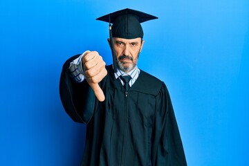 Middle age hispanic man wearing graduation cap and ceremony robe looking unhappy and angry showing rejection and negative with thumbs down gesture. bad expression.
