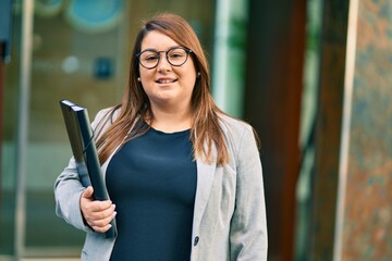 Young hispanic plus size businesswoman smiling happy holding binder at the city.
