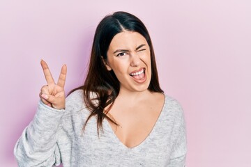 Young hispanic woman wearing casual clothes smiling with happy face winking at the camera doing victory sign. number two.