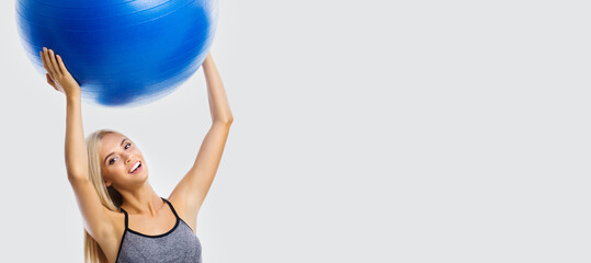 Portrait of beautiful smiling woman in sportswear holding fitness blue ball, isolated over grey color background. Blond girl in fit exercising studio concept. Wide banner composition image.