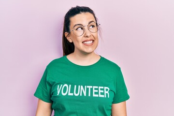 Young hispanic woman wearing volunteer t shirt smiling looking to the side and staring away thinking.