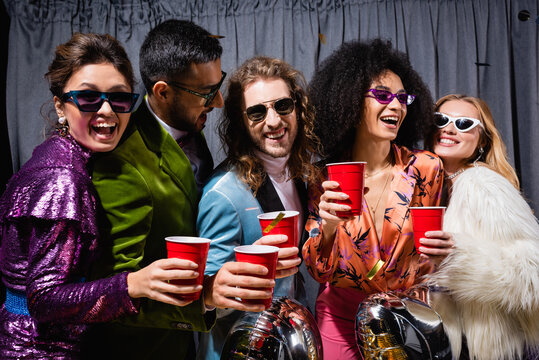 happy interracial friends in sunglasses celebrating with plastic cups on grey background.