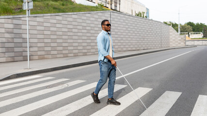 Young black visually impaired man wearing dark glasses, walking across city street, using cane - obrazy, fototapety, plakaty