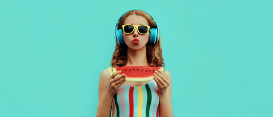 Obraz premium Summer fashion portrait of young woman in headphones listening to music with juicy slice of watermelon, female model blowing her lips posing on a colorful blue background