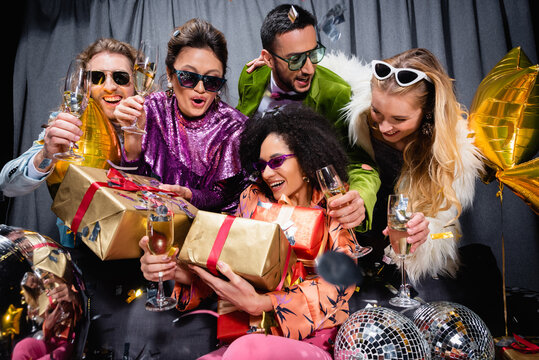 stylish african american woman with gift boxes sitting among interracial friends on grey background.