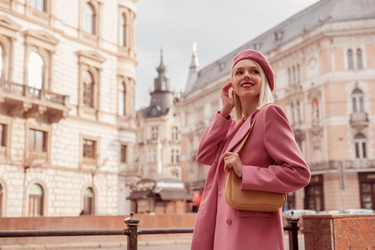 Elegant smiling woman wearing stylish pink blazer, beret, with yellow shoulder bag, posing in street of European city. Copy, empty space for text
