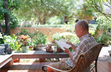 senior asian retired man sitting on bench and reading book at home backyard during free time