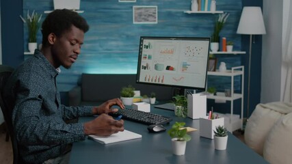 African american guy working from home, taking notes on notepad while remote working on computer from living room. Computer user using business internet online web communication job