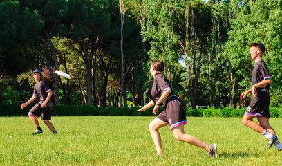 Group of young teenagers people in team wear playing a frisbee game in park oudoors. man tosses a frisbee to a teammate in an ultimate frisbee match. milennials friends outside in a garden having fun - obrazy, fototapety, plakaty