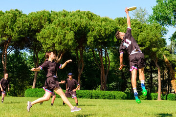 Group of young teenagers people in team wear playing a frisbee game in park oudoors. jumping man catch a frisbee to a teammate in an ultimate frisbee match. milennials friends outside in a garden - obrazy, fototapety, plakaty