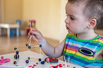 A little boy sculpts from plasticine at the table at home. Makes a tower of toothpicks and balls.