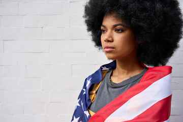 Proud confident patriotic African American gen z girl wrapped in usa flag looking in future with pride standing on white wall background. Equality and freedom in united states concept. - Powered by Adobe