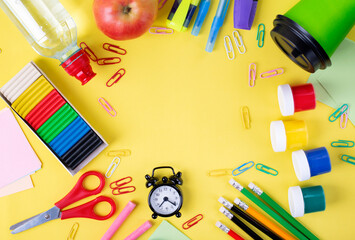 Back to school flat lay framing copy space. Set of stationery supplies, apple, water bottle, paper cup and alarm clock on yellow background. Colorful composition