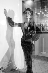Young fashionable brunette lady with hairstyle in long black dress with glittering diamonds posing sad in fitting room with large mirrors - 437727114