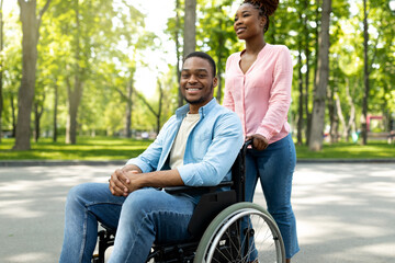 Happy black woman taking her impaired boyfriend in wheelchair for walk at city park