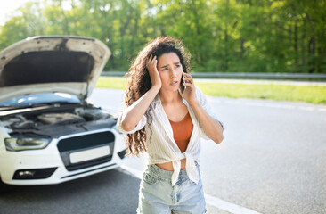 Millennial Caucasian woman with smartphone calling car towing service on roadside, standing near her broken vehicle