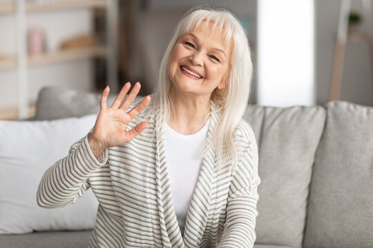 Mature woman waving at camera and smiling, sitting on sofa