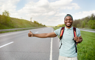 Smiling black guy with backpack flagging down car, catching free ride, hitchhiking on highway, free...