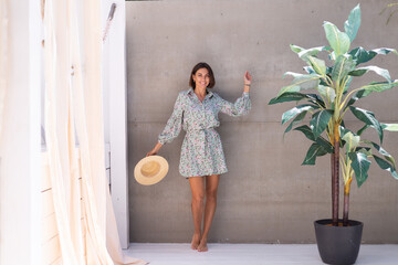 Beautiful woman in summer dress and straw hat by palm tree and gray wall on background, playful...