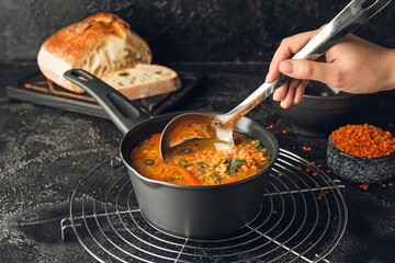 Female hand with saucepan of hot lentil soup and ladle on dark background