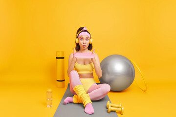 Impressed Asian sporty woman in activewear stretches arms with resistance band poses on fitness mat does exercises for losing weight increase flexibility looks shocked at camera isolated on yellow