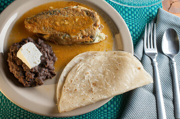 A stuffed Mexican chile with Mexican rice and beans on a plate, cuttlery on the side on a grey napkin
