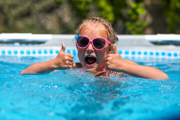 Naklejka premium Cheerful little girl smiling in sunglasses in the pool on a sunny day. The child showing thumbs up like gesture looking camera . Summer vacation