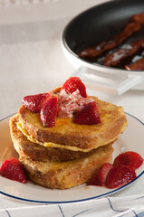 front view, close up of three layered French toast with sliced strawberries and and cream filling and a frying pan with bacon in background