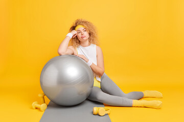 Losing weight keeping fit and pilates at home. Bored thoughtful sporty woman leans at fitness ball has workout or training dressed in activewear surrounded by sport equipment yellow background
