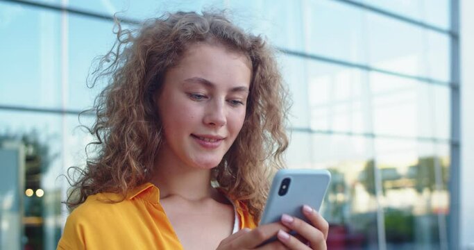 Caucasian young joyful woman with curly hair standing in city and sending message on smartphone smiling in good mood. Beautiful female surfing internet on mobile phone online. Close up concept