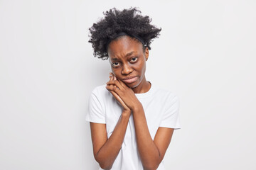 Fototapeta na wymiar Upset dejected dark skinned woman looks with miserable expression keeps hands near face wants to cry feels lonely wears casual t shirt isolated over white background. Depressed Afro teenage girl