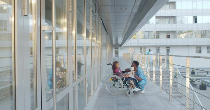 Laughing little girl sitting on wheelchair playing with nurse outside hospital