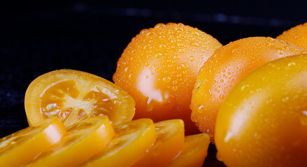 Beautiful whole yellow tomatoes with drops of water on the peel and a cut tomato on a black background.