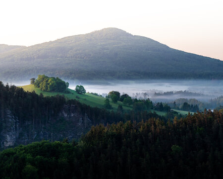 Mountain with fog in the valley and hill with trees during sunrise viewed from Mariina sk&aacute;la (Marienfels), Bohemian Switzerland, Czechia