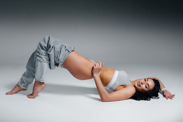 Obraz premium Young sexy brunette posing lying down in underwear and jeans on a gray background. The perfect athletic figure.