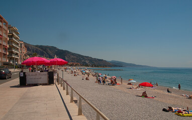 People spending time of a sunny beach in Menton, France. Red parasols. Clear blue sky and turquoise ocean.