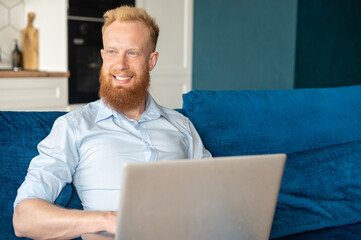 Cheerful charismatic young redhead man using laptop computer for studying or working online, freelance guy develops new project working remotely, happy bearded hipster guy looks aside and dreaming