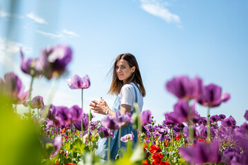Brunette in poppies field. She looks melancholic. Red and purple poppy flowers. Village life. White shirt. Sunny day. Blue sky.